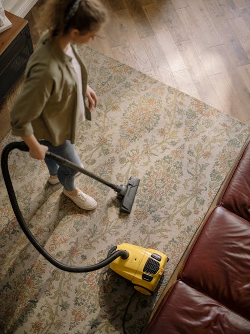 A professional cleaner dressed in a white protective suit, gloves, face mask, and blue overshoes uses a vacuum cleaner with a long hose to deep clean a beige carpet in a bright, minimalistic room. The