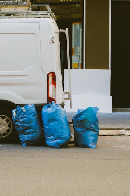 A blue waste bin is positioned outside the rear of a white, mobile cleaning service truck on a paved surface. The bin is made of plastic, with a hinged lid, and has visible dirt marks on its surface. 