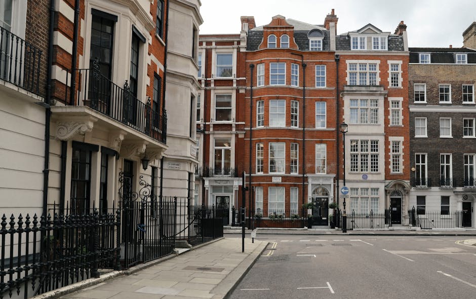 A multi-story red-brick residential building with decorative architectural details, featuring large white-framed sash windows, some with small balconies and black wrought-iron railings. The roof has m