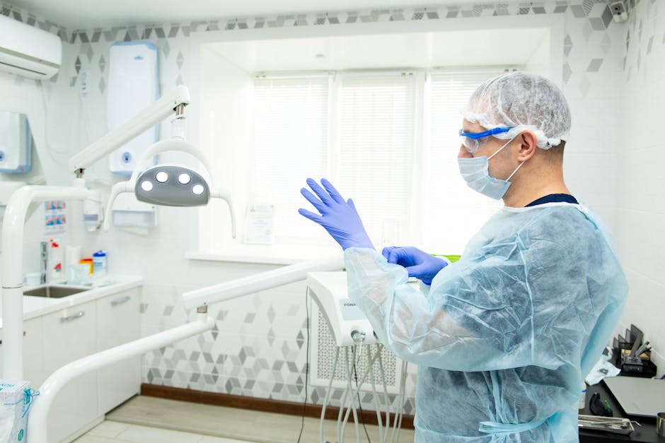 A healthcare professional dressed in light blue scrubs, a disposable gown, a face mask, protective glasses, and a hairnet, is preparing for surface cleaning in a clinical dental room. The room features white tiled walls with geometric patterns along the upper border, bright natural light coming through a window with horizontal blinds, and various dental tools and equipment neatly arranged on counters. The professional appears to be holding a cloth or disinfectant wipe, emphasizing hygienic and deep cleaning standards typical of a Harley Street clinic. The sterile setting, combined with the use of protective gear and cleaning supplies, highlights the emphasis on sanitisation and meticulous maintenance in medical environments, aligning with the high standards promoted by Carpet Cleaners W1 for their cleaning services in Marylebone, W1.