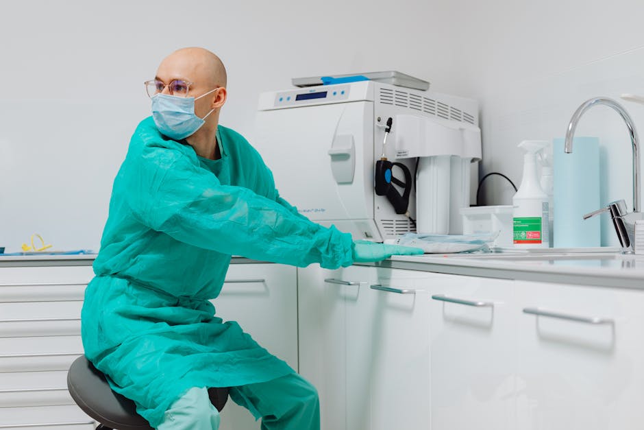A healthcare professional wearing a blue surgical mask, green medical scrubs, and gloves, seated on a stool in a clinical setting. The individual is reaching out with their right hand toward a white countertop with drawers, which holds medical equipment and cleaning supplies. On the countertop are a sterilisation device, a spray bottle, and other hygiene products. The room features clean, white surfaces, a stainless steel sink, and bright, even lighting, suggesting a sterile environment consistent with clinical cleaning standards. The background shows a plain white wall, emphasizing the cleanliness and hygiene typical of a medical or clinic space where disinfecting and surface cleaning are prioritized, reflecting high-level hygiene protocols managed by Carpet Cleaners W1 in line with Harley Street clinic cleaning standards.