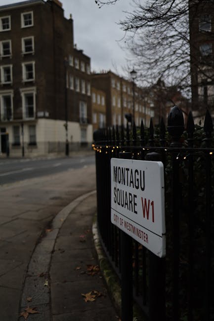 A street sign displaying 'Montagu Square W1' is mounted on a black wrought iron fence surrounding a small garden area. Fine string lights are affixed along the top of the fence. Behind the fence, leafless trees with sparse branches are visible, indicating a winter or late autumn season. In the background, rows of residential buildings with brick facades and multiple windows line the street, which appears to be relatively quiet and empty. The overcast sky casts a subdued, diffused light over the scene, emphasizing the urban environment of Marylebone High Street, W1. The overall atmosphere is calm, with a focus on the street sign and surrounding urban details, suitable for a guide on local area features or street-level visuals.