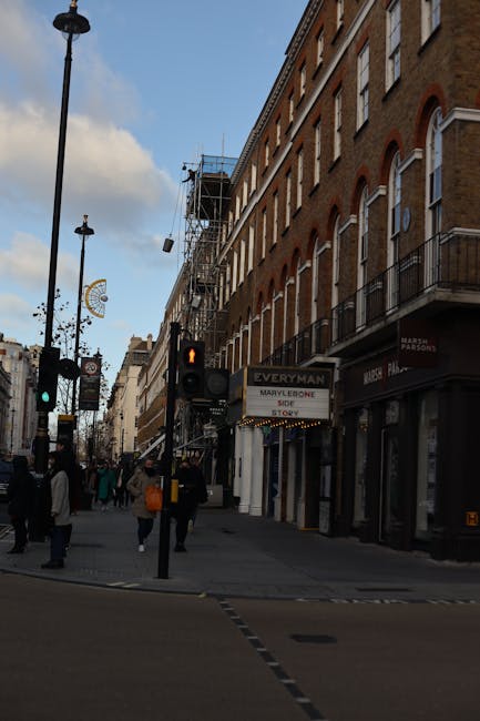 A street sign displaying 'Montagu Square W1' is mounted on a black wrought iron fence surrounding a small garden area. Fine string lights are affixed along the top of the fence. Behind the fence, leafless trees with sparse branches are visible, indicating a winter or late autumn season. In the background, rows of residential buildings with brick facades and multiple windows line the street, which appears to be relatively quiet and empty. The overcast sky casts a subdued, diffused light over the scene, emphasizing the urban environment of Marylebone High Street, W1. The overall atmosphere is calm, with a focus on the street sign and surrounding urban details, suitable for a guide on local area features or street-level visuals.
